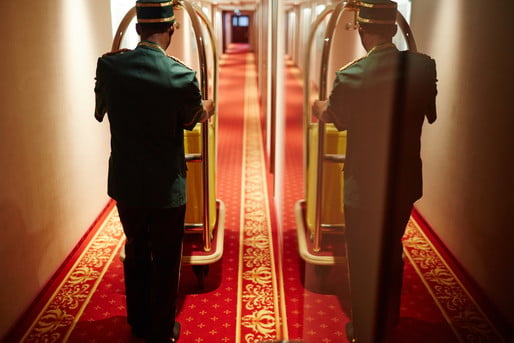 Back view portrait with mirror reflection of bellboy pushing luggage cart in hotel hallway, delivering bags to rooms