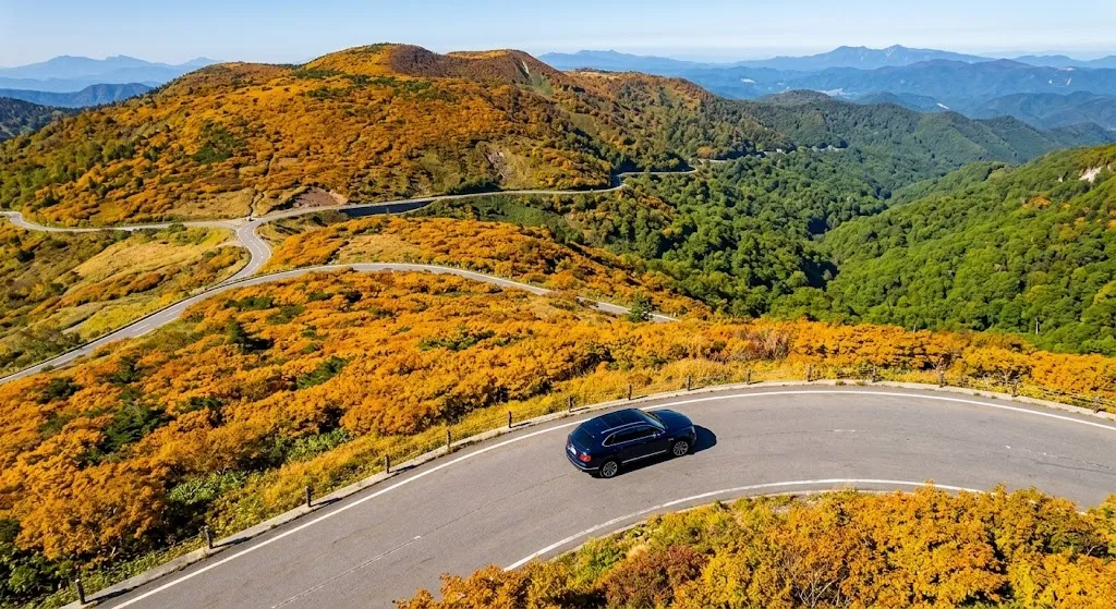 A luxury vehicle driving on a winding mountain pass in northern Japan, where high-altitude autumn golds contrast sharply with the deep green forests of the lower valleys.