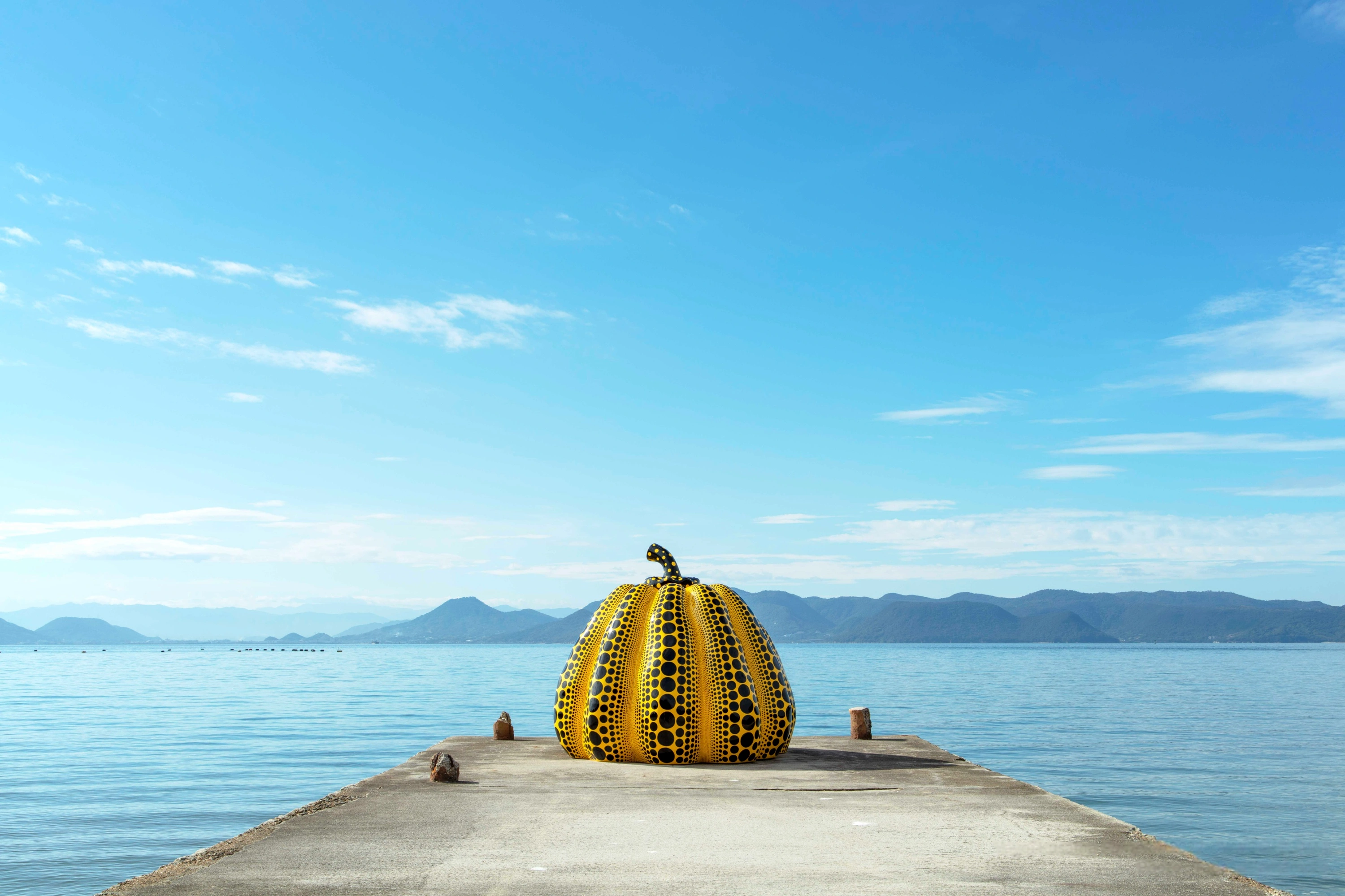 Large yellow and black spotted sculpture on a wooden pier extending over calm blue water with distant mountains on the horizon.